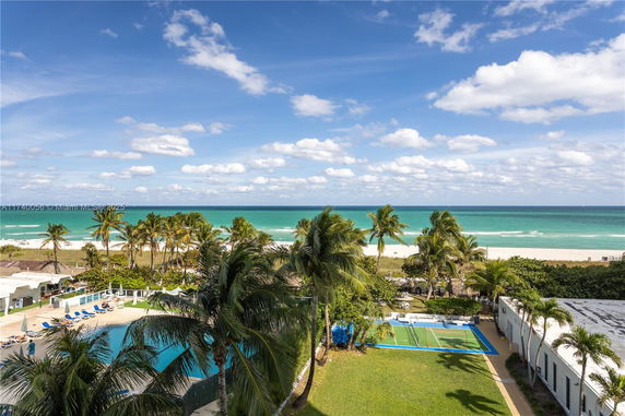 Wide angle view of a beach and ocean with palm trees, swimming pool, and a sports court.