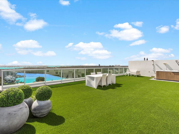 Rooftop with artificial grass, dining area, and glass railing overlooking water and cityscape.