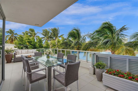 Outdoor patio area with glass table and wicker chairs overlooking palm trees and water.