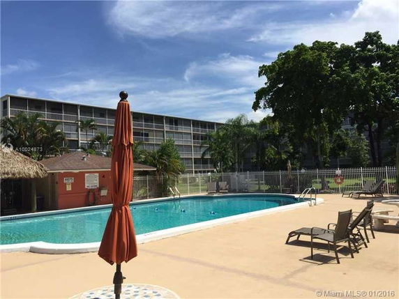 View of a multi-story residential building with balconies, next to a swimming pool and patio with lounge chairs.