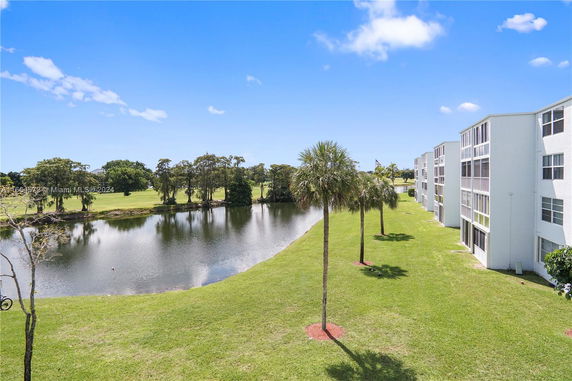 Exterior view of apartment buildings near a lake with trees and grassy area.