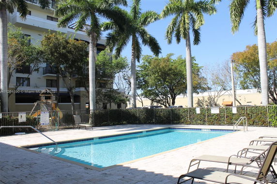 Outdoor swimming pool with lounging chairs surrounded by palm trees and a fence.