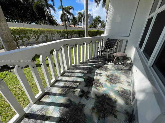 Balcony area with decorative railing, chair, table, and view of grassy area and palm trees.