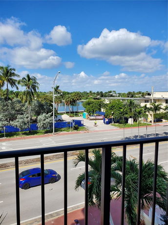 Panoramic view from a balcony showing a street, palm trees, and water in the distance under a partly cloudy sky.