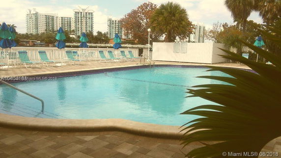 View of a swimming pool area surrounded by lounge chairs and umbrellas, with buildings visible in the background.