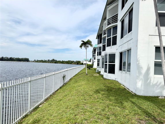 Side view of a building with large windows facing a body of water, with a grassy area and a white fence.