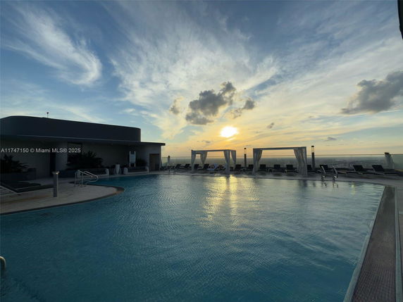 Rooftop pool area with lounge chairs and canopies overlooking a sunset.