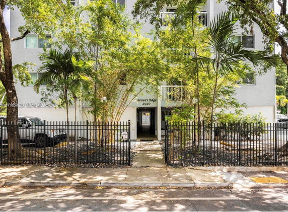 Front view of a multi-story residential building behind a metal fence with trees and plants.