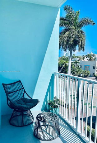 Balcony view with a chair and table, overlooking palm trees and nearby buildings.