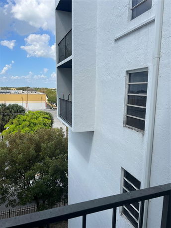 View of the side of a building with a balcony and windows, overlooking trees and other buildings in the distance.