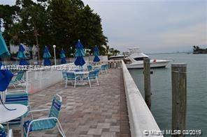 Outdoor seating area by the water with tables and blue umbrellas, adjacent to a waterway with a moored boat.