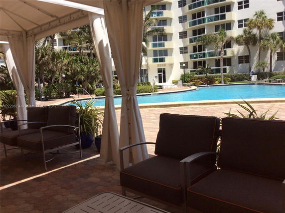 View from a shaded area with seating, looking out towards a swimming pool and apartment building.