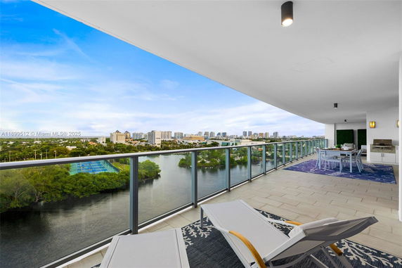 View from a spacious balcony overlooking a cityscape and waterway, with lounge chairs and outdoor dining area.