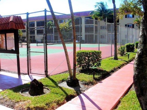 Tennis court surrounded by a fence, with trees and a walkway.