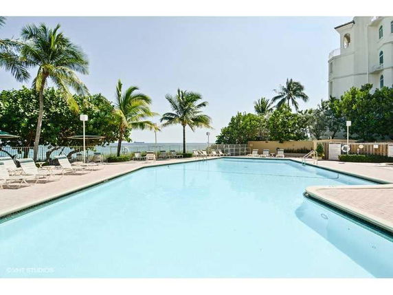 View of a swimming pool area with palm trees and a building in the background.