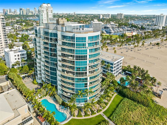 View of a modern, multi-story residential building with glass balconies and a swimming pool area. The building is located near a beach with volleyball courts.