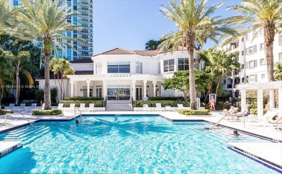 View of a large swimming pool in front of a multi-story building with palm trees.
