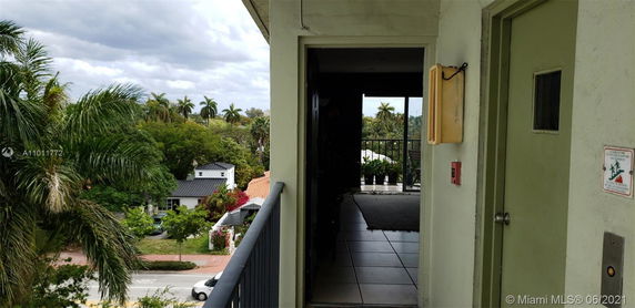 Partial view of a building entrance with vegetation and neighboring houses in the background.