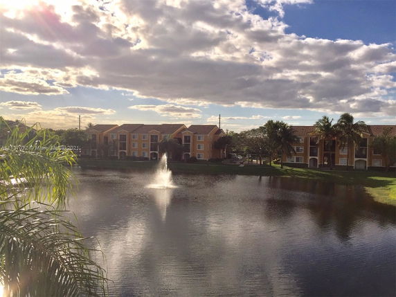 View of apartment buildings by a pond with a fountain, surrounded by trees and open sky.