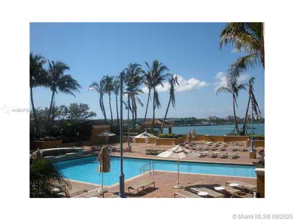 View of a swimming pool area with palm trees and a body of water in the background.