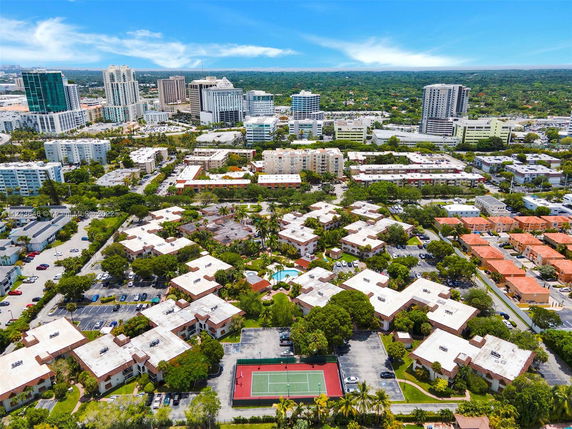Aerial panoramic view of a residential area with multiple buildings and a tennis court, surrounded by trees and urban landscape.