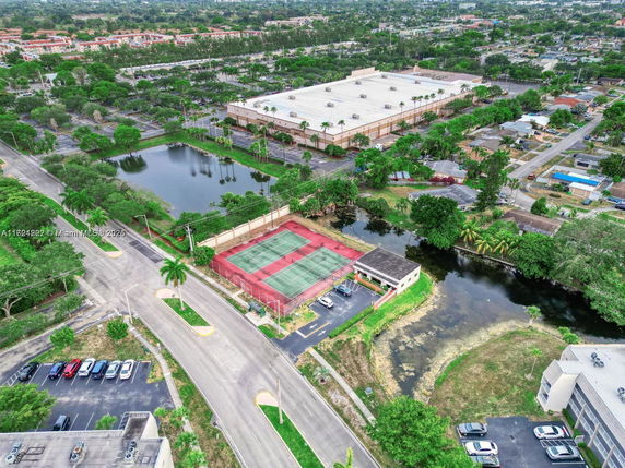 Aerial view of a tennis court, parking lot, and surrounding area with trees and water bodies.