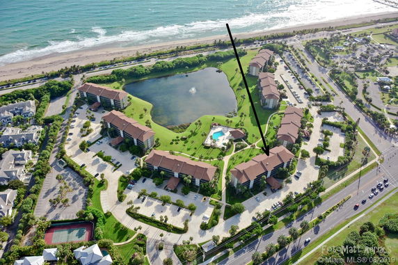 Panoramic view of a complex of multi-story residential buildings near the beach, with a central pond and swimming pool.