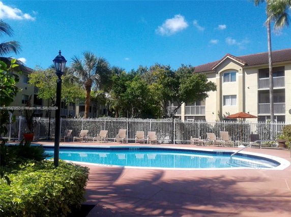 Outdoor swimming pool area with lounge chairs and a fence, surrounded by trees and apartment buildings in the background.