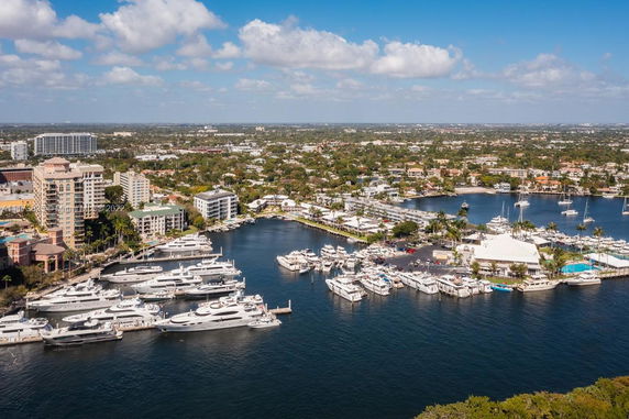 View of a marina with multiple boats docked along the waterfront and various buildings in the background.