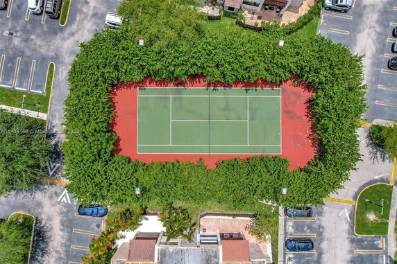 Aerial view of a tennis court surrounded by greenery with adjacent parking areas.