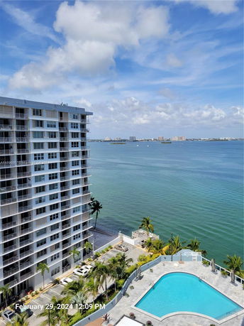 View of a high-rise building near a large body of water with a swimming pool and parking area in the foreground.