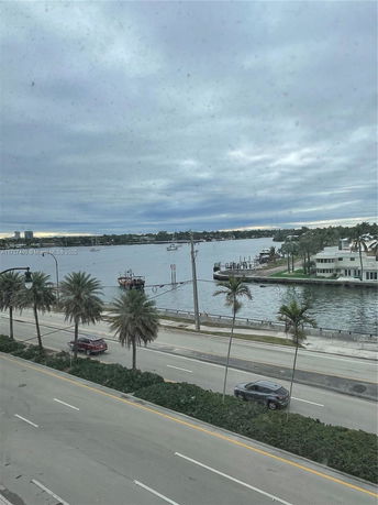 View of a road and waterfront area with palm trees and buildings across the water.