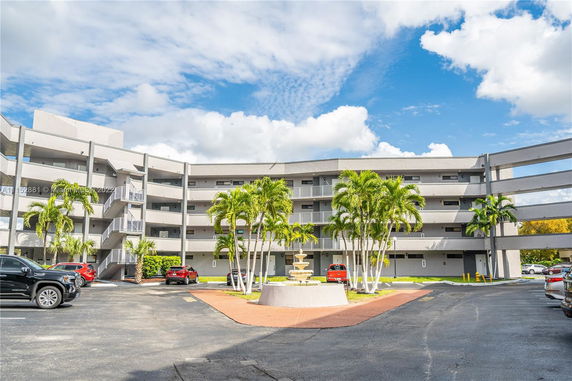 Front view of a multi-story apartment building with balconies and a central courtyard featuring palm trees and a fountain.