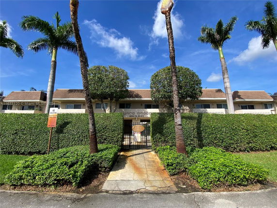 Front view of a two-story residential building with a hedge and palm trees.