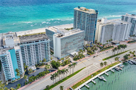 Aerial view of high-rise beachfront buildings near a road with palm trees, boats, and ocean.