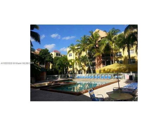 Outdoor swimming pool area with surrounding palm trees and multi-story buildings.