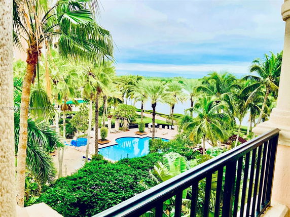 View from a building balcony overlooking a tropical landscape with palm trees and a blue swimming pool.