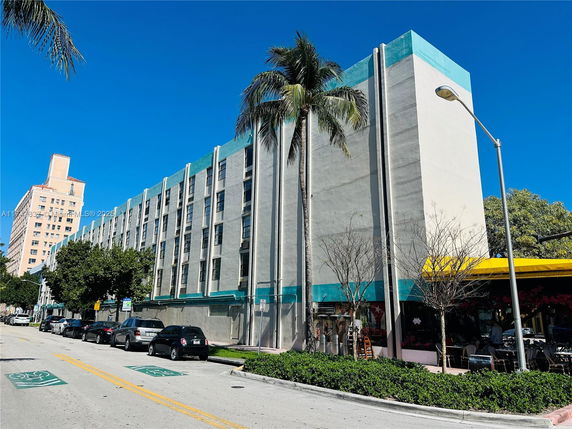 Front view of a multi-story building with a teal and gray color scheme, featuring palm trees and street parking.