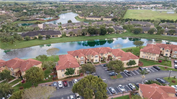 Aerial panoramic view of a residential area with red-roofed houses, a parking lot, and a water body surrounded by greenery.