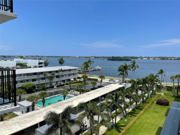 View of the surrounding area from a building, including other buildings, a swimming pool, and a water body with palm trees.