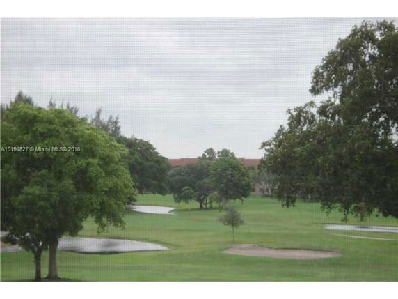 View of a green field with scattered trees and water bodies under a cloudy sky.