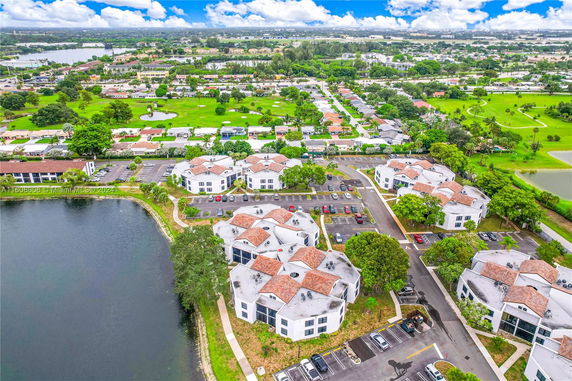 Aerial view of a residential complex with several white buildings featuring red-tiled roofs, surrounded by green areas, parking lots, and water bodies.