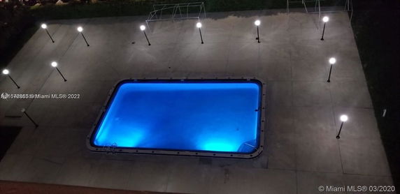 Aerial view of a rectangular swimming pool illuminated at night with surrounding pavement and light posts.