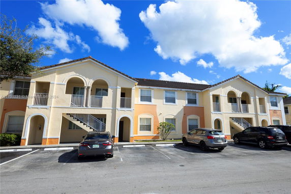 Front view of a two-story apartment building with arched balconies and multiple parking spaces.