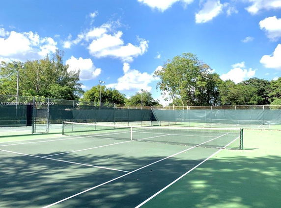 Image of a tennis court with green surfaces and surrounding trees under a clear sky.