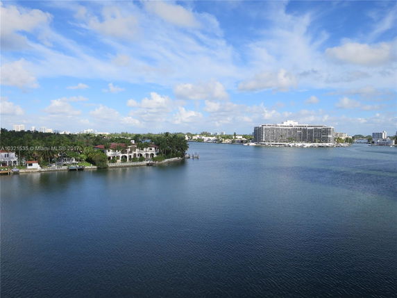 View of a large waterfront building surrounded by water, with smaller residential houses in the distance.