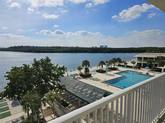 Elevated view of a swimming pool and surrounding recreational area next to a body of water, with trees and distant buildings across the water.