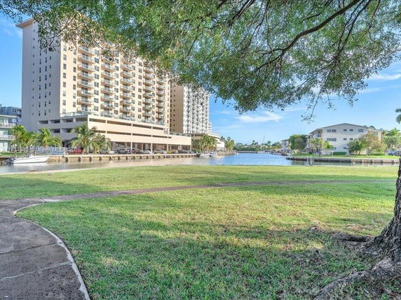 View of a tall residential building by a waterway, surrounded by grassy area and trees.