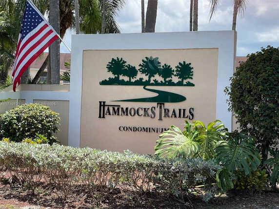 Entrance sign for Hammocks Trails Condominium with surrounding greenery and an American flag.