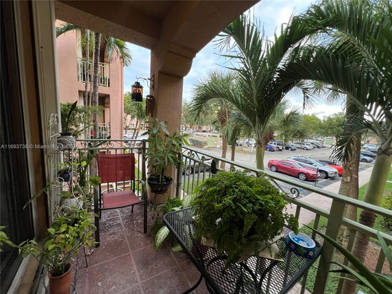 View from a balcony with plants and chairs, overlooking a parking area and palm trees.